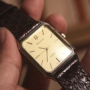 Puede incluir: Un reloj Bulova Quartz dorado con una esfera rectangular y una correa de cuero marrón. La esfera del reloj tiene agujas e indicadores dorados. La correa es de color marrón oscuro con un patrón texturizado similar al de un cocodrilo.