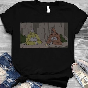 Peut inclure: T-shirt noir avec une illustration de dessin animé de deux créatures ressemblant à des Bigfoot assises en méditation. L'une est jaune, l'autre est marron. L'arrière-plan représente un paysage rocheux.