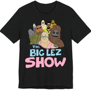 以下が含まれることがあります： 黒いTシャツ。漫画のキャラクターのグラフィックが特徴です。「THE BIG LEZ SHOW」の文字が青とピンクで表示されています。デザインには、サスカッチのような生き物や、その他風変わりなキャラクターなど、さまざまなアニメーションフィギュアが含まれています。