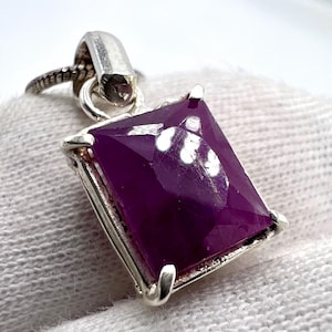 Puede incluir: Colgante de plata con una gran gema púrpura rectangular facetada. La gema está engastada en un marco de plata y sujeta a una fianza y cadena de plata. El colgante se muestra sobre un fondo blanco.