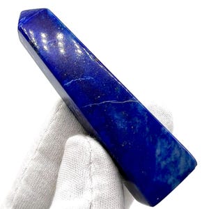 May include: A polished, rectangular lapis lazuli crystal obelisk. The deep blue stone has white and gold flecks and a crack running along its surface. The obelisk is held by a gloved hand.