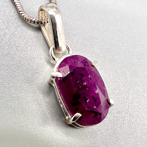 Puede incluir: Un collar con colgante de plata con una piedra preciosa ovalada de color magenta intenso. La piedra preciosa está engastada en un bisel de plata y tiene inclusiones visibles. La cadena es una cadena de plata de serpiente.