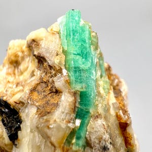 以下が含まれることがあります： 鮮やかな緑色のエメラルド結晶のクローズアップ。 エメラルドが焦点で、結晶構造をしています。 周囲のマトリックスはベージュ、茶色、白の鉱物の混合物で、自然なコントラストを生み出しています。