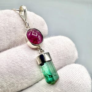 May include: A silver pendant necklace featuring a vibrant pink oval gemstone above a green tourmaline crystal. The pendant hangs from a delicate silver chain, showcasing the natural beauty of the gemstones.