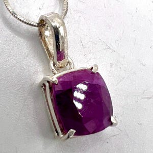 May include: A silver pendant with a large, faceted, deep magenta gemstone. The gemstone is square-shaped and set in a silver bezel. The pendant hangs from a silver chain, creating a classic jewelry piece.