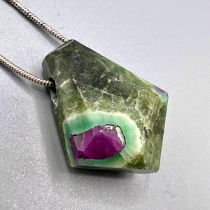 May include: A green and purple gemstone pendant with a silver chain. The pendant has a rough, natural texture with a vibrant purple center and a green border. The silver chain is thin and delicate.