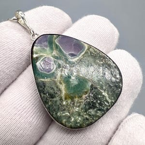 Puede incluir: Colgante de piedra preciosa con cadena de plata. El colgante presenta una piedra grande de forma irregular con una mezcla de colores verdes, morados y blancos. La piedra tiene una superficie pulida y está engastada en un bisel de plata.