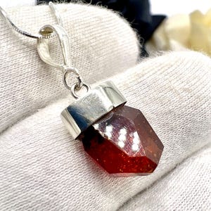 Może przedstawiać: Fasetowany, czerwonobrązowy wisiorek z kamieniem szlachetnym ze srebrną oprawą i łańcuszkiem. Wisiorek ma geometryczny kształt i odbija światło. Łańcuszek jest cienki i delikatny, odpowiedni do codziennego noszenia.