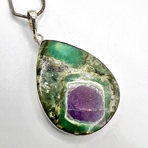 May include: A teardrop-shaped pendant with a silver bail and chain. The pendant features a large, polished stone with a central purple section surrounded by a white border and a green and gray mottled exterior. The stone is set in a silver bezel.