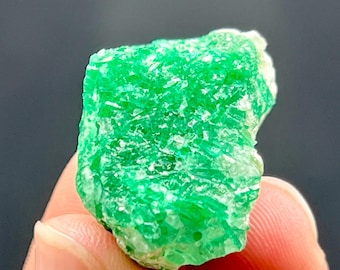 May include: A vibrant green emerald crystal with a rough, textured surface. The gemstone is held in a person's hand, showcasing its natural, unpolished form. The emerald's color is a rich, vivid green, with areas of lighter green and white.