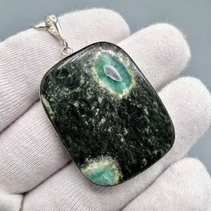 May include: A rectangular pendant with a dark green and black stone, featuring teal and purple accents. The pendant is attached to a silver chain and bail. The stone has a textured appearance.