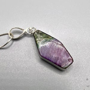 Puede incluir: Un collar de plata con un colgante de forma alargada inusual. El colgante muestra una mezcla de tonos morados, verdes y blancos, engastado en un marco plateado. La cadena del collar también es plateada.