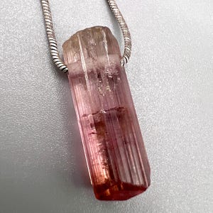 May include: A pink tourmaline crystal pendant with a silver chain. The crystal is a long, rectangular shape with a natural, textured surface. The color transitions from a light pink to a deeper red at the base. The silver chain is a snake chain style.