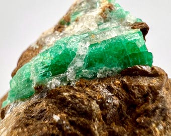 May include: Close-up of a raw emerald crystal cluster. The vibrant green emerald crystals are set against a matrix of brown rock. The emerald crystals have a translucent appearance, with visible internal structures.