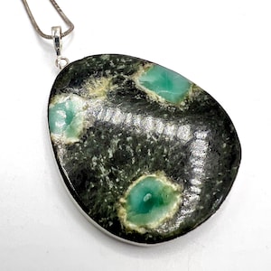 以下が含まれることがあります： 楕円形の石を使ったペンダントネックレス。濃い緑色のベースに、薄い緑色とターコイズ色のパッチがあります。石は銀色の金属フレームにセットされ、細い銀色のチェーンから吊り下げられています。