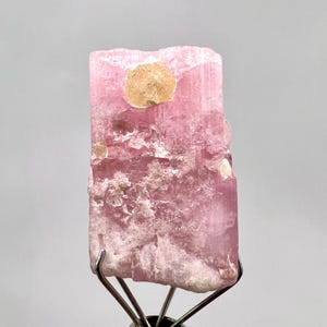 May include: A raw, pink tourmaline crystal specimen. The crystal has a rectangular shape with a rough, textured surface. A patch of tan-colored material is visible on the front. The crystal is mounted on a metal stand, showcasing its natural beauty.
