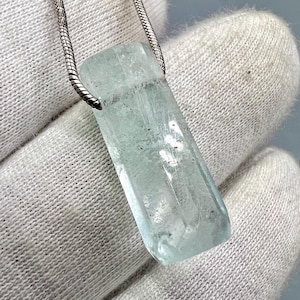 Könnte beinhalten: Ein rechteckiger, hellblauer Aquamarin-Anhänger an einer Silberkette. Der Edelstein ist durchscheinend mit feinen inneren Einschlüssen. Die Kette ist aus feinem, strukturiertem Silber. Der Anhänger befindet sich vor einem weißen Stoffhintergrund.