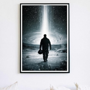 May include: Black and white framed art print. A silhouetted figure walks away from the viewer, holding a helmet, towards a bright light emanating from a swirling galaxy. The artwork is displayed on a white wall above pillows.