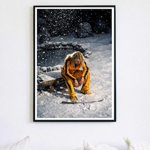 May include: A framed print depicts a person in a yellow jumpsuit with black stripes, seated on a wooden bench in a snowy outdoor scene. A katana lies in the snow. The image is set against a backdrop of falling snow and a dark, blurred background.