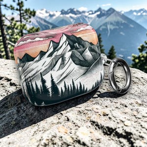 May include: A white and black mountain range design on a protective case. The case features a sunset sky with pink and orange hues. A silver carabiner clip is attached to the case. The case is designed for earbuds.