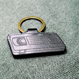 May include: A black leather keyring shaped like a rectangular VHS cassette tape with a debossed graphic of the tape reels and casing details, attached to a circular gold-toned metal split ring for keys.