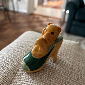 May include: A ceramic figurine of a light brown teddy bear lying in a high-heeled shoe. The shoe is painted with a green vamp and a yellow heel. The bear is curled up, with its paws near its face.