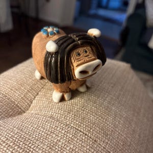 May include: A whimsical ceramic figurine of a Highland cow. The cow has a brown body, black fringe, and white accents on its horns and feet. A blue and white floral design adorns its back. The figurine is a decorative item.
