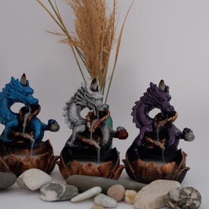 May include: Three dragon incense burners in blue, silver, and purple. Each dragon sits atop a lotus flower base, with incense smoke visible. Decorative stones and dried grass are in the foreground.