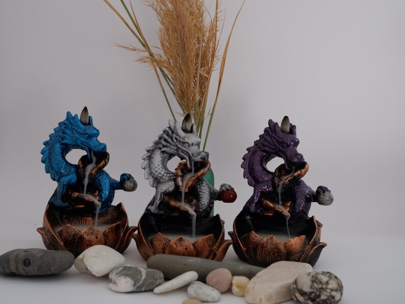 May include: Three dragon incense burners in blue, silver, and purple. Each dragon sits atop a lotus flower base, with incense smoke visible. Decorative stones and dried grass are in the foreground.