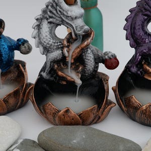 May include: Three dragon incense burners in blue, white, and purple. Each dragon sits atop a lotus flower base with a bronze finish. Incense smoke flows from the dragon's mouth, creating a mystical effect. Decorative home decor.