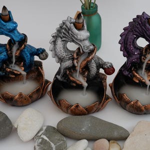 May include: Three dragon-shaped incense burners in blue, gray, and purple. Each dragon sits atop a lotus flower base with a copper finish. Incense smoke flows from the dragon's mouth. Decorative stones are in the foreground.