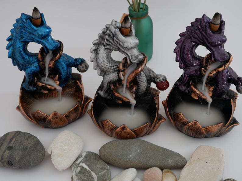 May include: Three dragon-shaped incense burners in blue, gray, and purple. Each dragon sits atop a lotus flower base with a copper finish. Incense smoke flows from the dragon's mouth. Decorative stones are in the foreground.