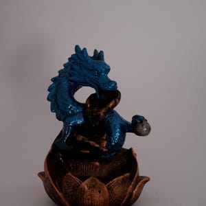 May include: A blue dragon figurine with a textured finish, perched atop a bronze-colored lotus flower base. The dragon is detailed with a metallic sheen, and the base has a detailed petal design. The figurine is set against a plain background.