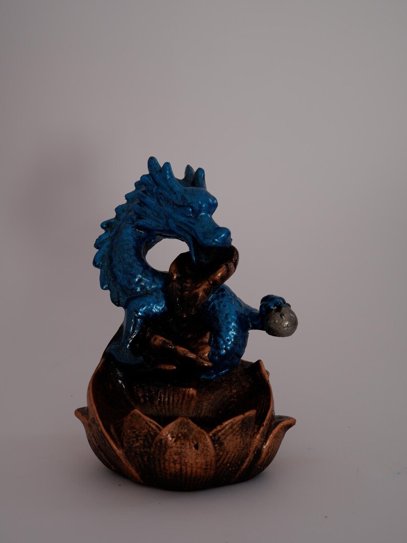 May include: A blue dragon figurine with a textured finish, perched atop a bronze-colored lotus flower base. The dragon is detailed with a metallic sheen, and the base has a detailed petal design. The figurine is set against a plain background.