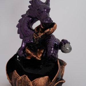 May include: A purple dragon figurine with bronze and black accents. The dragon is perched atop a lotus flower base. The dragon is holding a silver orb. The figurine is a decorative item.