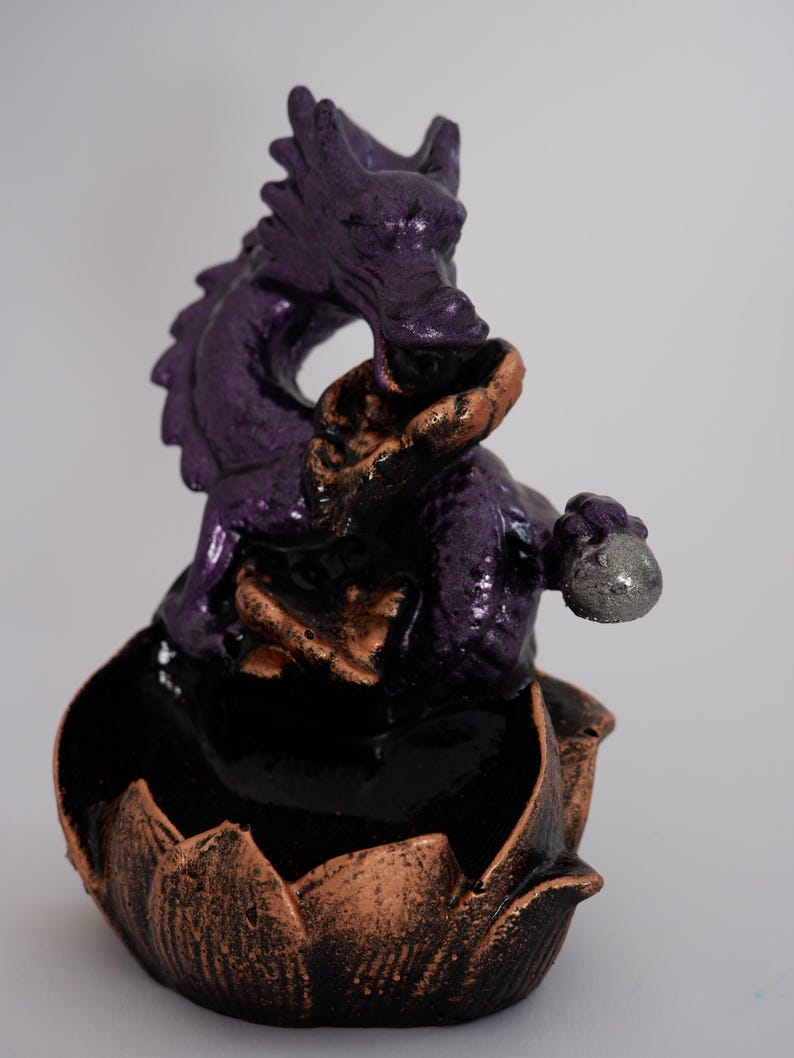May include: A purple dragon figurine with bronze and black accents. The dragon is perched atop a lotus flower base. The dragon is holding a silver orb. The figurine is a decorative item.