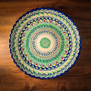 Könnte beinhalten: Ein großer, runder Keramikteller mit gewelltem Rand und einem komplexen, handbemalten radialen Muster. Das Design besteht aus konzentrischen Ringen mit blauen, grünen, gelben und roten Blumen- und geometrischen Motiven auf weißem Grund.