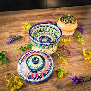 Könnte beinhalten: Eine Keramikschale und ein Deckel mit einem lebendigen Blumenmuster in Blau, Rot, Gelb und Grün. Die Schale steht auf einem Holzuntersetzer, und ein kleiner Räuchergefäßbrenner befindet sich in der Nähe. Dekorative Blumen und Grünzeug umgeben die Gegenstände auf einer Holzoberfläche.