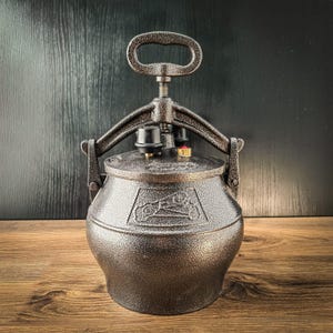 Op de afbeelding: Een donkerbronskleurige metalen snelkookpan met een getextureerde, gehamerde afwerking. Het heeft een grote handgreep aan de bovenkant, een vergrendelingsmechanisme met twee zijarmen en een deksel met een zwart ventiel en een rode knop.