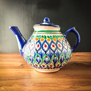 Könnte beinhalten: Eine Keramik-Teekanne mit einem handgemalten Muster aus blauen, grünen und orangefarbenen Rautenmotiven. Die Teekanne hat einen bauchigen Körper, einen geschwungenen Henkel, einen spitzen Ausguss und einen passenden Deckel mit Knauf.