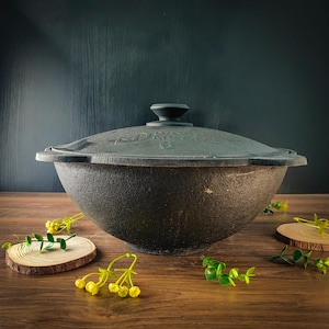 Puede incluir: Una olla de cocina de hierro fundido negro con tapa a juego. La olla tiene forma redondeada y superficie texturizada. La tapa tiene un asa en la parte superior. La olla está sobre una superficie de madera.