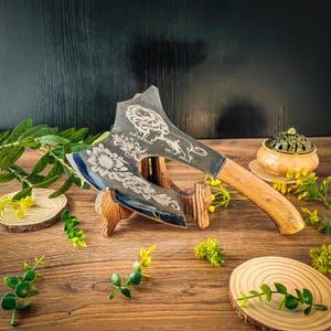 Könnte beinhalten: Eine dekorative Axt mit Holzgriff und Metallklinge mit eingravierten Blumen- und Löwenmotiven. Die Axt wird auf einem Holzständer präsentiert, mit einem kleinen hölzernen Räuchergefäß und Grünzeug im Hintergrund.