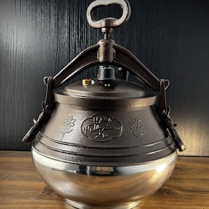 May include: A dark brown cast metal pressure cooker with a textured finish, featuring a polished silver-tone base, a top locking handle mechanism, and embossed floral designs on the sides with Arabic script text centred on the lid.
