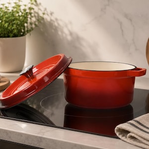Puede incluir: Una olla redonda de hierro fundido esmaltado en color rojo brillante con una tapa a juego que incluye un pomo negro. La olla tiene dos asas laterales y un interior de color crema, apoyada sobre una superficie de cocina de vidrio negro brillante.