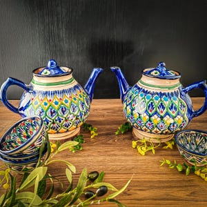 Può includere: Due teiere in ceramica e due piccole ciotole con intricati motivi blu, verdi, gialli e rossi. Le teiere hanno manici e beccucci blu e poggiano su sottobicchieri di legno. Rami di ulivo e fiori gialli sono disposti intorno alle teiere e alle ciotole.