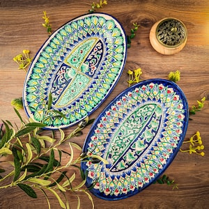 Può includere: Due piatti ovali in ceramica con disegni intricati e colorati. I piatti presentano un bordo blu e un motivo centrale in verde, giallo e rosso. Un piccolo contenitore di legno con un coperchio decorativo in metallo è nelle vicinanze, insieme a rametti di verde.