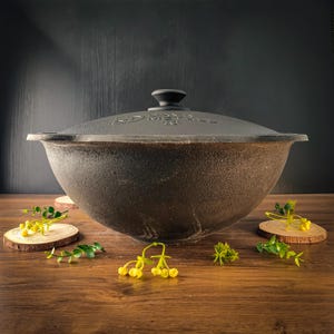 Puede incluir: Una olla grande de hierro fundido de color gris oscuro con una tapa a juego. La olla se encuentra sobre una superficie de madera, con rodajas de madera decorativas y flores amarillas. La tapa tiene un pequeño asa y texto.