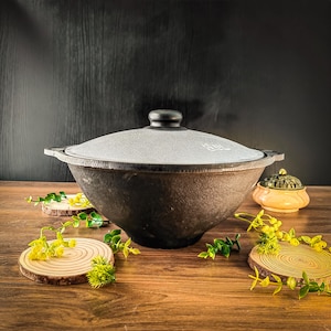 May include: A large, dark brown cast iron pot with a grey lid and black handle. The pot sits on a wooden surface, surrounded by decorative wooden coasters and greenery. A small, decorative incense burner is also present.