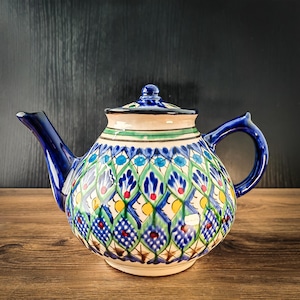 Può includere: Una teiera in ceramica con base color crema e un motivo a rombi dipinto a mano nei toni del blu, verde e giallo. La teiera include un coperchio abbinato con un piccolo pomello, un manico curvo e un beccuccio affusolato, rifiniti in blu.