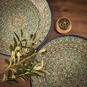 May include: Two large, round, decorative plates with a teal and floral design, and a blue rim. A small wooden incense holder with a decorative metal top sits between the plates. A sprig of green leaves and olives is in the foreground.